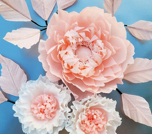 Elegantly crafted large paper peony in soft blush tones with feathered edges and a trio of white, detailed paper flowers, each showcasing a complex layering of petals, presented against a calming blue backdrop that enhances the visual texture and handcraft
