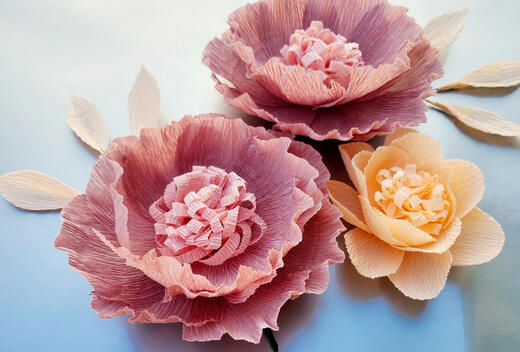 An array of handcrafted paper flowers, featuring rich mauve and soft peach hues, with realistic leaf accents, all artfully arranged on a tranquil blue canvas.