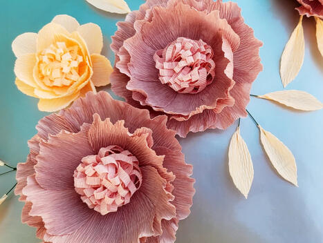 Three dimensional paper flowers in deep mauve with woven centers and peach counterparts, set against a pastel blue backdrop, showcasing exquisite artistry.