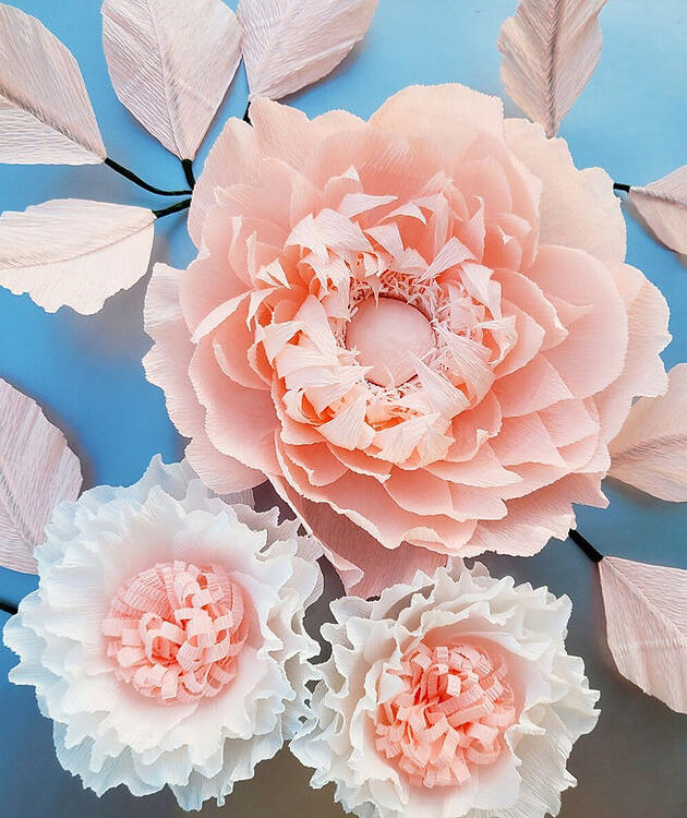 Artificially crafted, large blush pink peony and smaller white flowers with meticulously arranged paper petals displayed on a light blue backdrop, highlighting the delicate craftsmanship and realistic floral design.